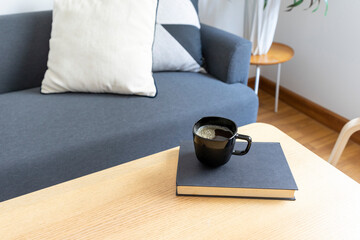 Wall Mural - Books and black cup of coffee on round wooden table in sofa background, cozy lifestyle concept. Scandinavian interior.