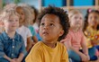 © imagineRbc - A diverse group of toddlers sit in a classroom, looking attentively up and to the left. The child in the center is wearing a yellow shirt