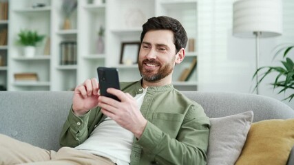 Happy handsome man using mobile phone lying on cozy sofa in living room at home. Smiling male browses social networks, chats online, watches something interesting, writes reads messages on smartphone