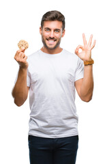 Wall Mural - Young handsome man eating chocolate chips cookie over isolated background doing ok sign with fingers, excellent symbol