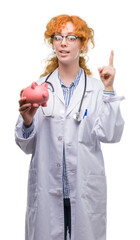 Wall Mural - Young redhead doctor woman holding piggy bank surprised with an idea or question pointing finger with happy face, number one