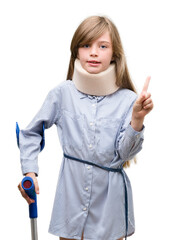 Sticker - Young blonde child injured wearing neck collar and crutches surprised with an idea or question pointing finger with happy face, number one