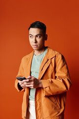 A handsome Asian man in a stylish orange jacket holding a cell phone against an orange background in a studio setting.