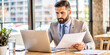© hobonski - Focused Businessman Reviewing Documents at Office Desk. A serious businessman in a suit intently reviews documents while working on his laptop in a bright, modern office.