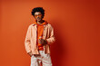 © LIGHTFIELD STUDIOS - Stylish young African American man posing confidently in trendy orange jacket and white pants on a vibrant orange backdrop.