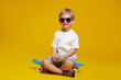 © Davidovici - Adorable little kid wearing casual outfit and sunglasses smiling while sitting on skateboard with cross legged against yellow background.