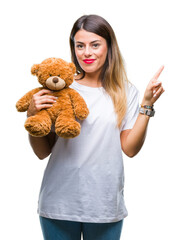 Poster - Young beautiful woman holding teddy bear plush over isolated background very happy pointing with hand and finger to the side