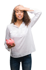 Canvas Print - Young hispanic woman holding piggy bank stressed with hand on head, shocked with shame and surprise face, angry and frustrated. Fear and upset for mistake.