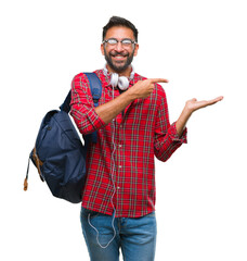 Wall Mural - Adult hispanic student man wearing headphones and backpack over isolated background amazed and smiling to the camera while presenting with hand and pointing with finger.