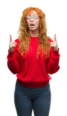 Canvas Print - Young redhead woman wearing red sweater amazed and surprised looking up and pointing with fingers and raised arms.