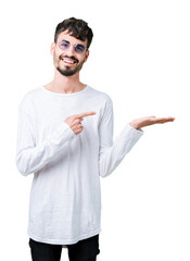 Canvas Print - Young handsome man wearing sunglasses over isolated background amazed and smiling to the camera while presenting with hand and pointing with finger.