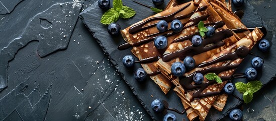 Fototapete - Chocolate crepes with blueberries sauce and mint leaves on a slate plate in a top view shot with a focused area left for text or design known as copy space image