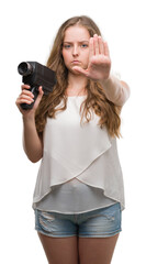 Canvas Print - Young blonde woman holding super 8 video camera with open hand doing stop sign with serious and confident expression, defense gesture