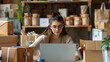 © narumon - Female entrepreneur managing her online store from a laptop She is surrounded by packaging, products, and shipping materials.