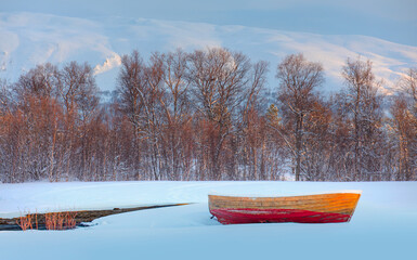 Wall Mural - orange wooden boat covered with layers of snow at sunset - cracks on the surface of the ice and dead