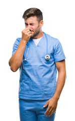 Canvas Print - Young handsome doctor surgeon man over isolated background smelling something stinky and disgusting, intolerable smell, holding breath with fingers on nose. Bad smells concept.