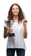 © Krakenimages.com - Young hispanic woman holding trophy surprised with an idea or question pointing finger with happy face, number one