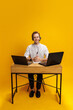 © master1305 - Portrait of smiling young man in white t-shirt, call center employee sitting at desk in headset, with two laptops against yellow background. Concept of customer support service, assistance, employment
