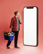 © master1305 - Thoughtful young man in checkered shirt, with basket of groceries looking at empty smartphone screen against pink background. Store website. Concept of online shopping, food, delivery service, app
