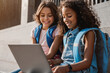 © InsideCreativeHouse - Two happy elementary middle school girls friends pupils classmates using laptop gadgets looking on screen doing homework project together sitting on the stairs steps outdoors
