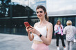 © BullRun - Young smiling Caucasian woman using a mobile app on smartphone while wearing workout attire, standing outdoors. earphones in ears, demonstrating use of mobile technology for fitness and connectivity
