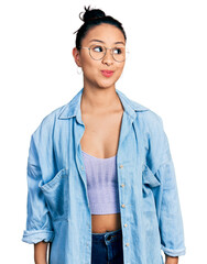 Beautiful hispanic woman wearing casual denim jacket and glasses smiling looking to the side and staring away thinking.