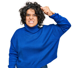 Canvas Print - Young hispanic woman with curly hair wearing turtleneck sweater angry and mad raising fist frustrated and furious while shouting with anger. rage and aggressive concept.