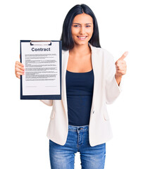 Sticker - Young beautiful latin girl holding clipboard with contract document smiling happy and positive, thumb up doing excellent and approval sign