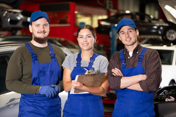 Wall Mural - Two young guys and young woman mechanics in uniform posing in car service station