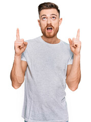 Wall Mural - Young redhead man wearing casual grey t shirt amazed and surprised looking up and pointing with fingers and raised arms.