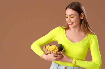 Poster - Beautiful young woman holding wicker basket with sweet mango fruits on brown background