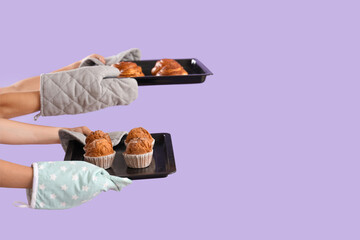 Wall Mural - Female hands in mittens holding baking trays with tasty pastry on purple background