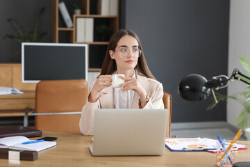 Wall Mural - Beautiful young businesswoman working with laptop and drinking coffee at table in office