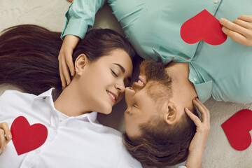 Wall Mural - Portrait of a young brunette man and woman with closed eyes lying on the floor with red hearts and smiling close. Happy couple in love. Care, tenderness and Valentines day concept.