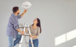 © Studio Romantic - Young man standing on the ladder changing light bulb in the room with his smiling wife. Happy couple installing the chandelier in their new empty apartment. Electricity replacing concept.