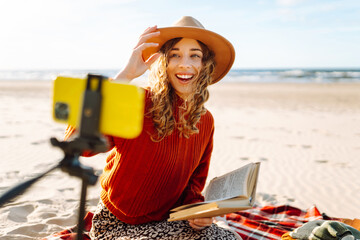 Wall Mural - A young woman sits on the beach and records a video for her subscribers. Happy woman creates content. Travel, blogging, technology concept