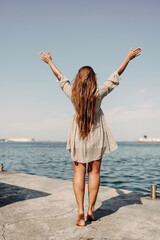 Wall Mural - woman in a dress stands on a dock by the water, her arms raised in the air. Concept of freedom and joy, as the woman is celebrating or expressing her happiness.