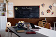 © Seventyfour - Background shot of empty primary school classroom with working desk, open laptops and student supplies. Study room decorated with blackboard and kids crafts hanging on beige wall, copy space