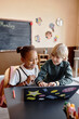 © Seventyfour - Vertical shot of two smiling primary students of black and white ethnic group studying together on laptop having fun in class, copy space