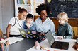 © Seventyfour - Diverse group of primary learners around female teacher of Black ethnicity explaining new information using laptop sitting at desk during elective lesson
