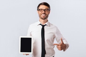 Poster - Look Here. Cheerful Businessman Holding Digital Tablet Pointing Finger At Blank Screen Over White Background. Mockup, Studio Shot