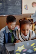 © Seventyfour - Vertical shot of two interested kids looking at laptop screen engaged in lesson with integrated educational technology, copy space