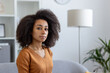 © Liubomir - Young woman with natural curly hair sitting on couch in modern living room. She appears thoughtful and calm, wearing mustard sweater. Background includes white shelves and standing lamp.