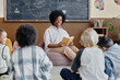 © Seventyfour - Female teacher of Black ethnicity in bean bag chair reading fairy tale aloud to attentive children sharing thoughts about story sitting around in classroom