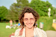© Connect Images - A smiling young woman with curly hair and glasses is standing outdoors on a sunny day, with blurred greenery and people in the background.