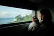 © Connect Images - A woman enjoys a warm beverage while gazing out a train window at a serene coastal landscape during a journey.