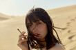 © Connect Images - A woman smoking a cigarette in the desert with windswept hair partially covering her face.