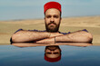 © Connect Images - Man with a beard wearing a red fez hat rests his chin on his crossed arms, reflected on a glossy surface against a desert backdrop.