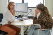 © Connect Images - Two women engaged in a serious conversation at an office workspace with a computer monitor in the background.