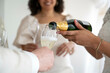 © Connect Images - Close-up of champagne being poured into a glass at a celebration with smiling guests in the background.